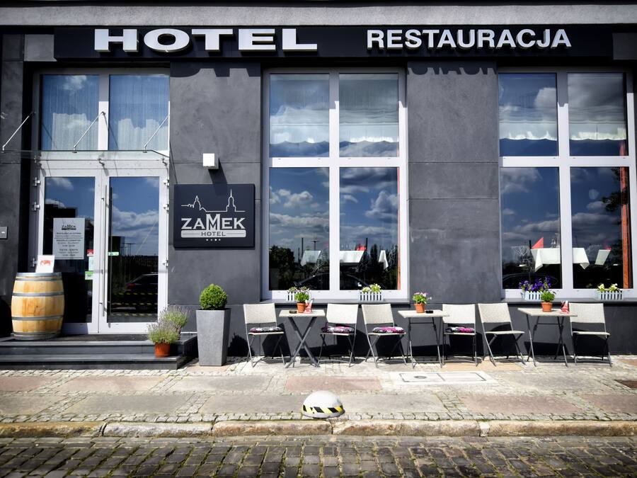 A hotel exterior with outdoor seating, a large window, and a sign reading "ZAMEK HOTEL RESTAURACJA".