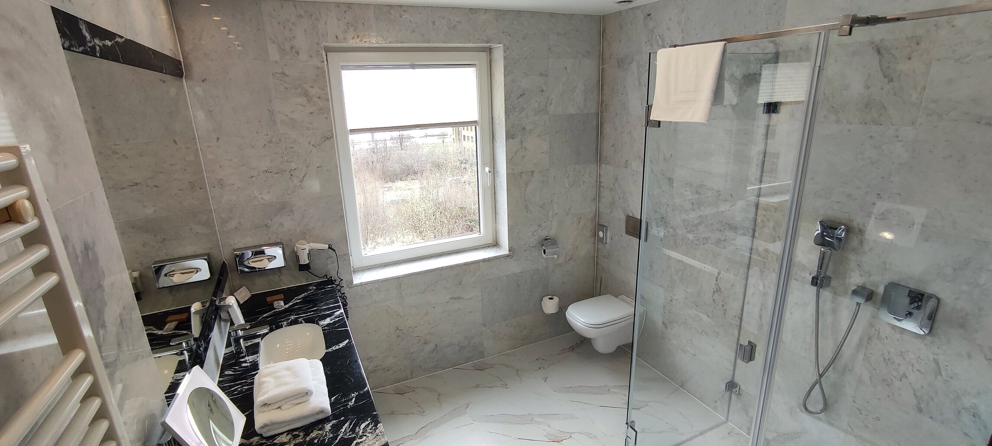 Une salle de bain dotée d’une douche à parois de verre, de toilettes, d’un lavabo avec un plan de toilette en marbre et d’une fenêtre laissant entrer la lumière naturelle.