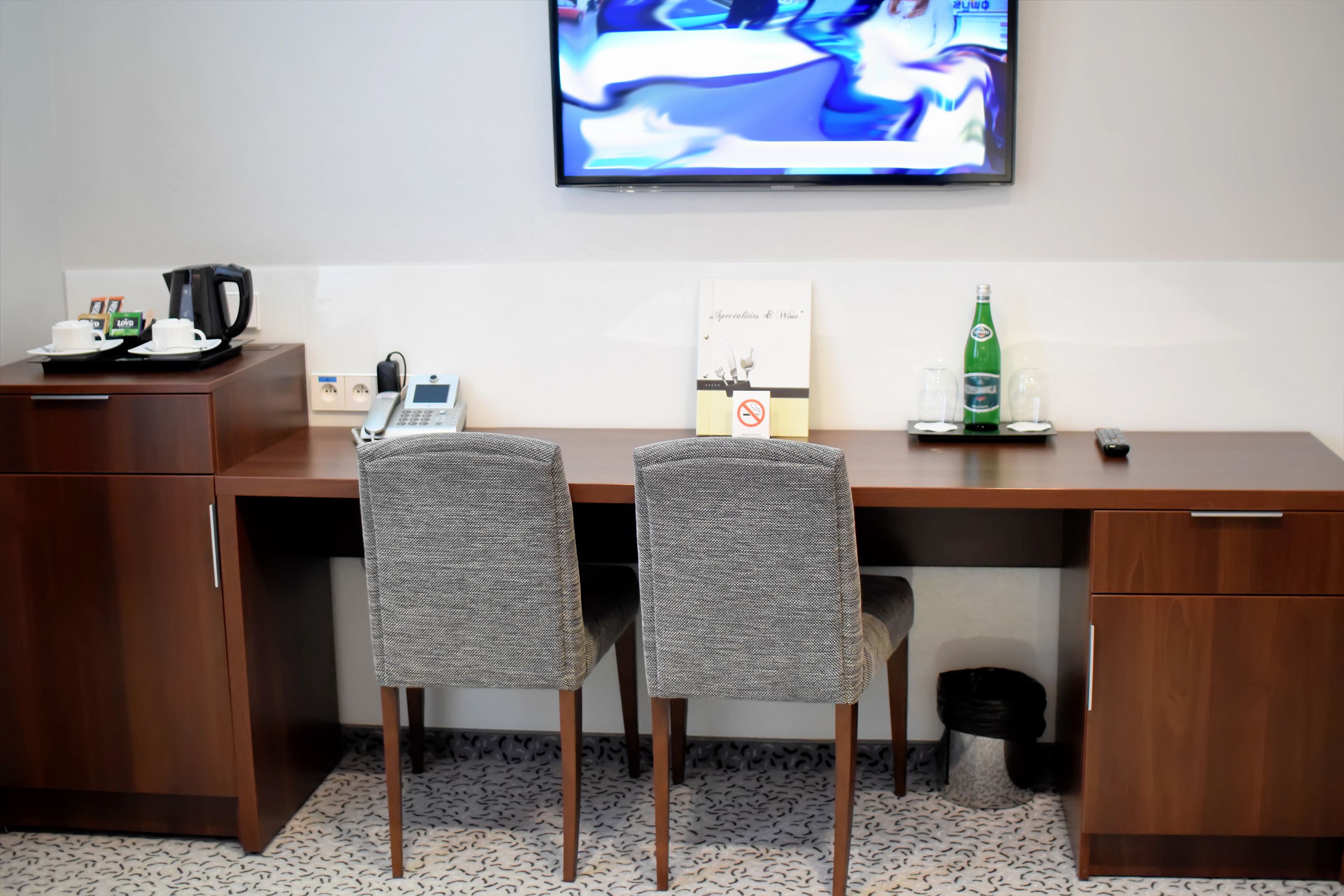 A hotel room with a wooden desk, two grey chairs, a coffee maker, a telephone, a television mounted on the wall, and a small table with a bottle and glasses.