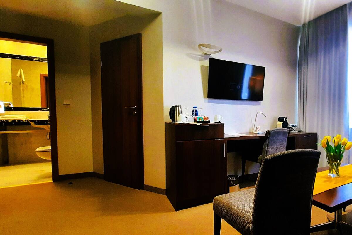A hotel room with a bed, a desk with a chair, a TV mounted on the wall, a window with curtains, and a small table with a vase of flowers.