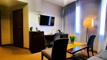 A hotel room with a bed, a desk with a chair, a TV mounted on the wall, a window with curtains, and a small table with a vase of flowers.