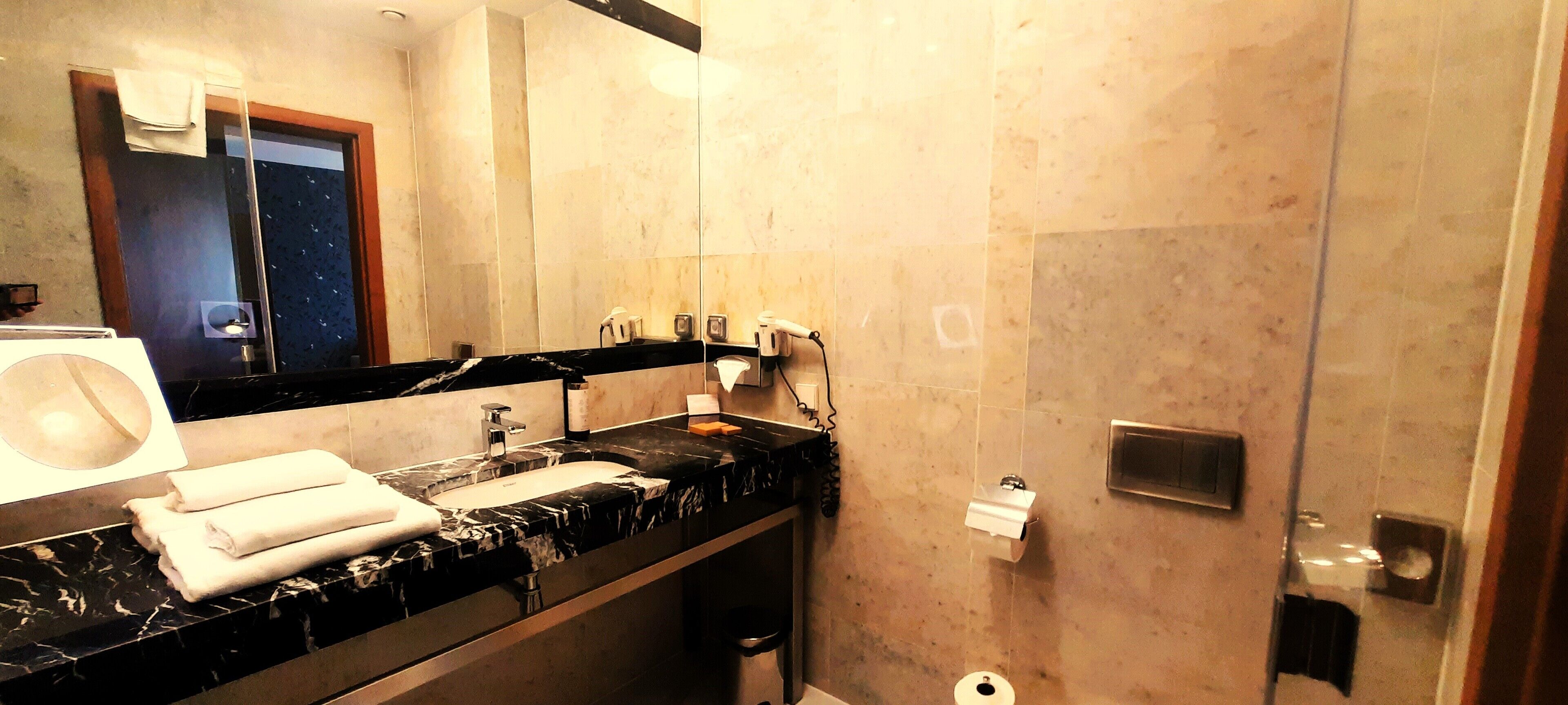 Une salle de bain moderne avec un comptoir en marbre noir, un lavabo et un miroir. On y trouve des serviettes sur le comptoir, un porte-rouleau de papier toilette et un espace douche doté d’une porte en verre.