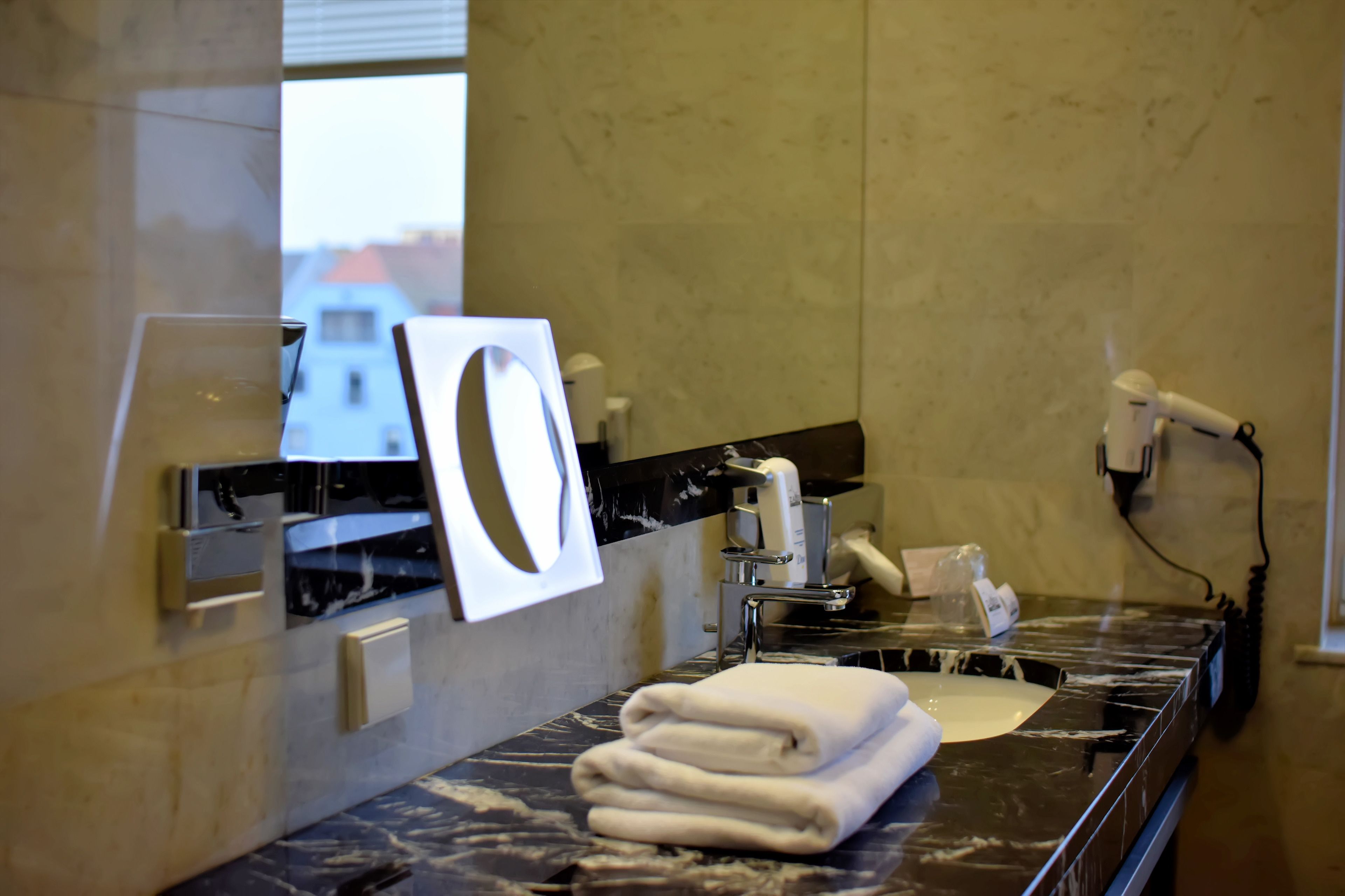 A bathroom with a marble countertop, a towel rack, a hand dryer, and a window with a view of buildings.