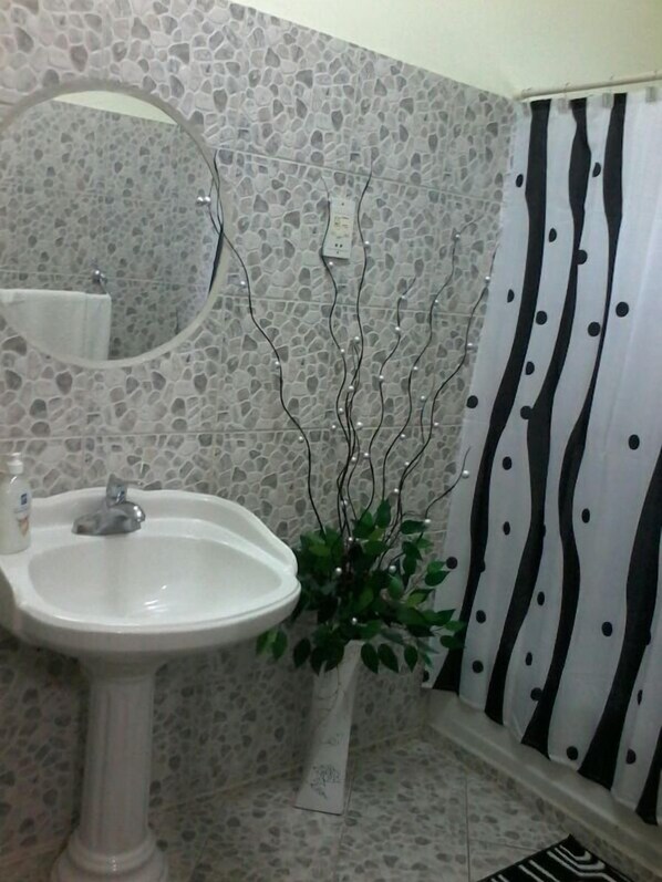 Baño con lavamanos blanco, espejo, planta decorativa y cortina de ducha con estampado en blanco y negro.