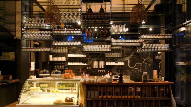 Una zona de bar con estantes llenos de vasos y botellas, una vitrina con pasteles y un mostrador con una caja registradora.