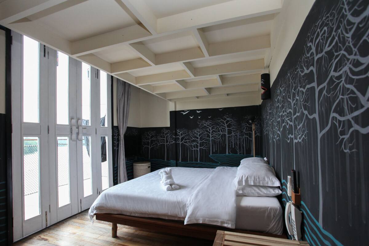 Une chambre avec un grand lit, une fenêtre avec des rideaux et un mur orné d’une fresque représentant un arbre.