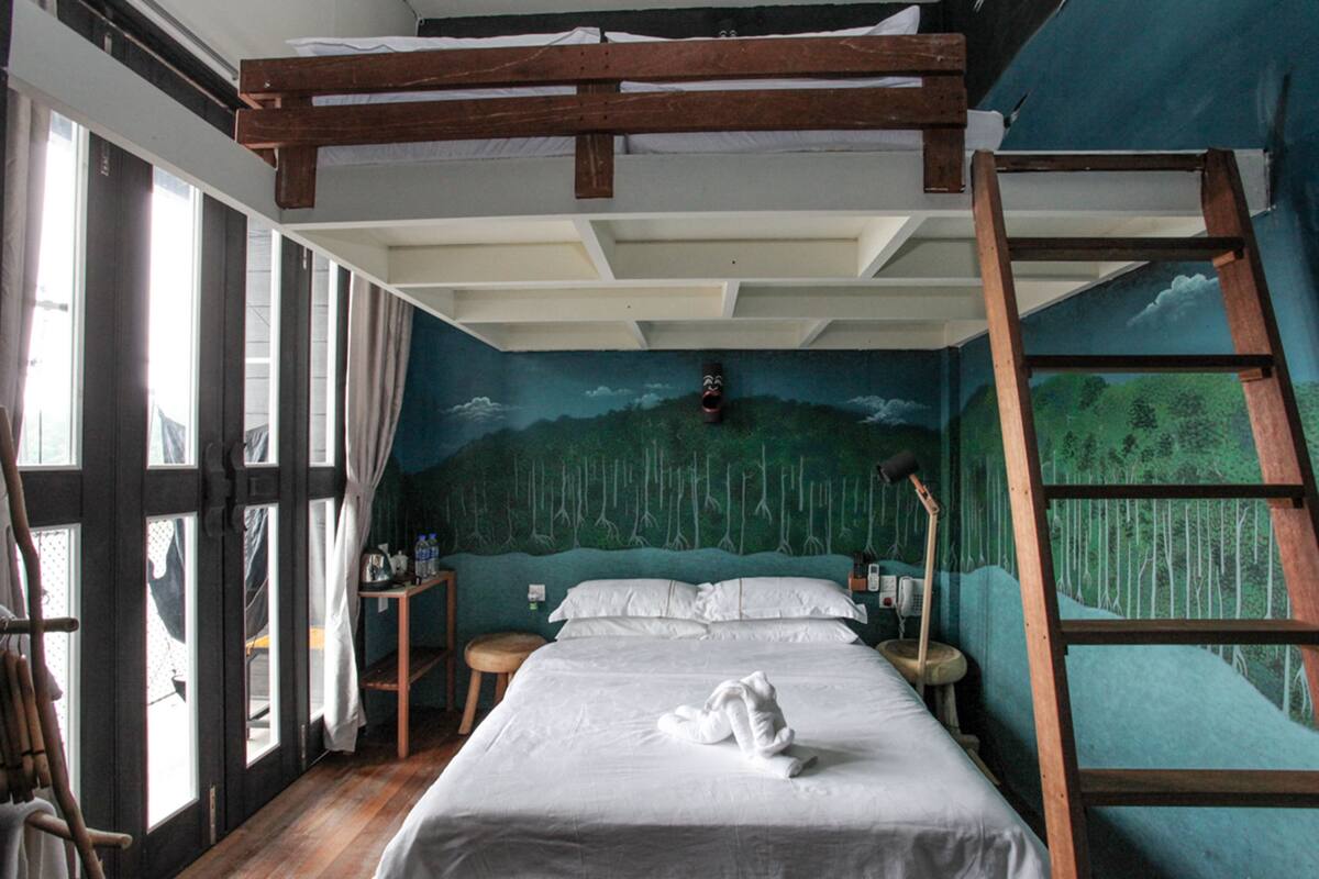 Une chambre avec un lit superposé, une échelle en bois, un bureau avec une chaise, un petit tabouret et une fresque représentant une forêt sur le mur.