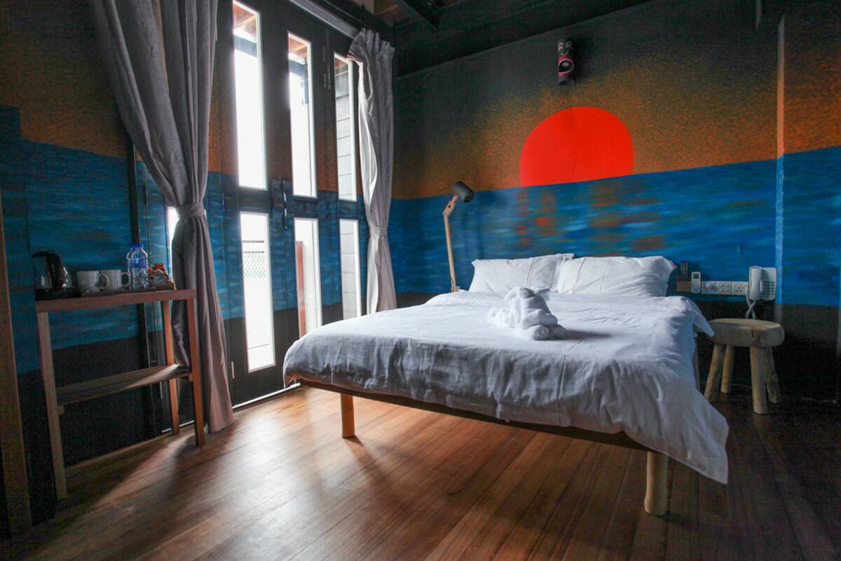Une chambre avec un lit en bois, une table de chevet, une lampe et une vue sur le coucher de soleil par la fenêtre.