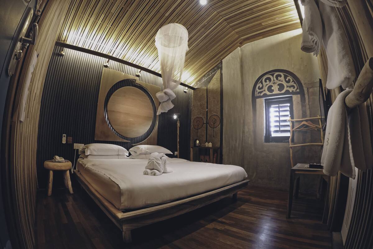 A bedroom with a large bed, a round mirror, a bedside table with a lamp, and a hanging light fixture.