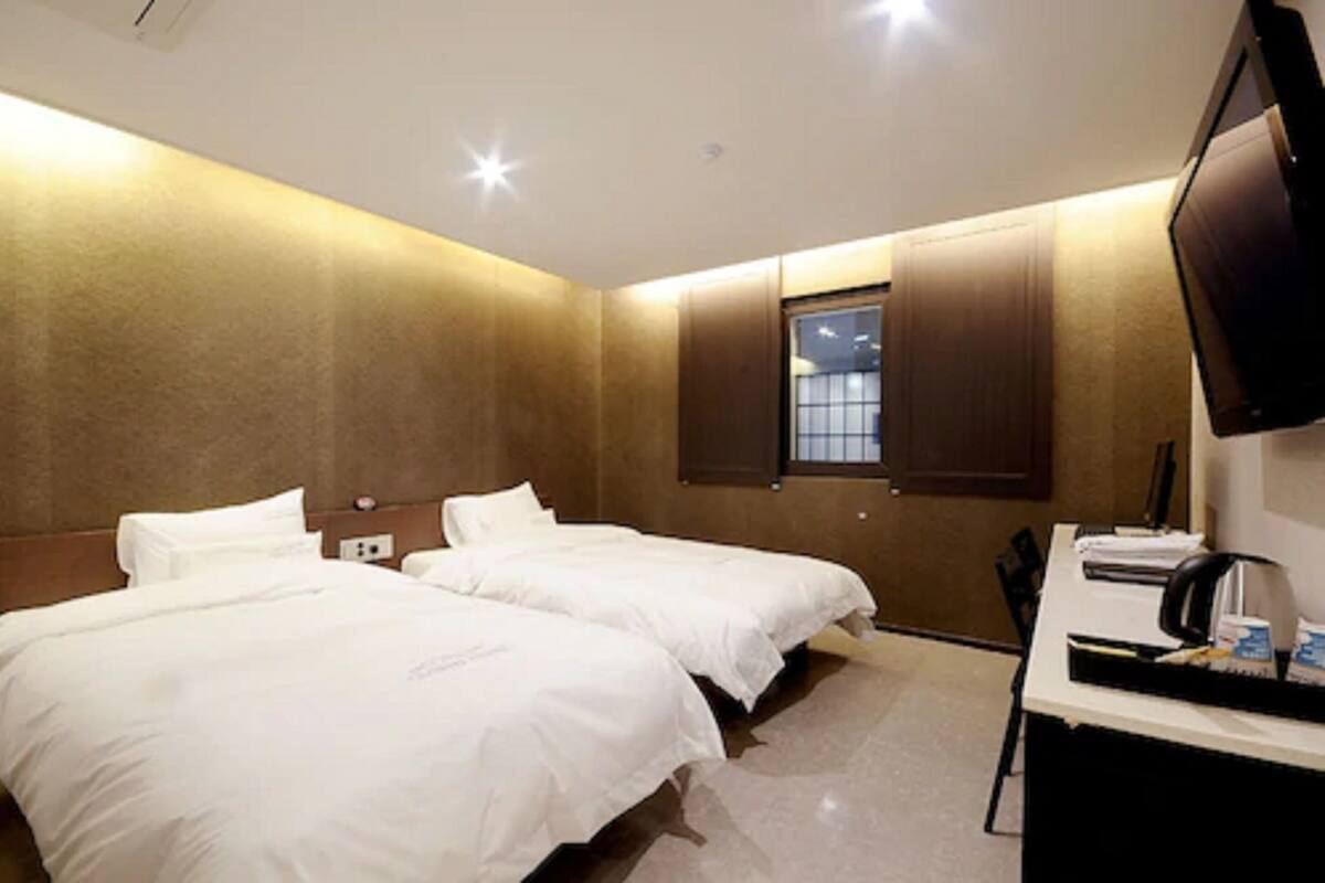 Quarto de hotel com duas camas, uma escrivaninha com computador e uma televisão fixada na parede.