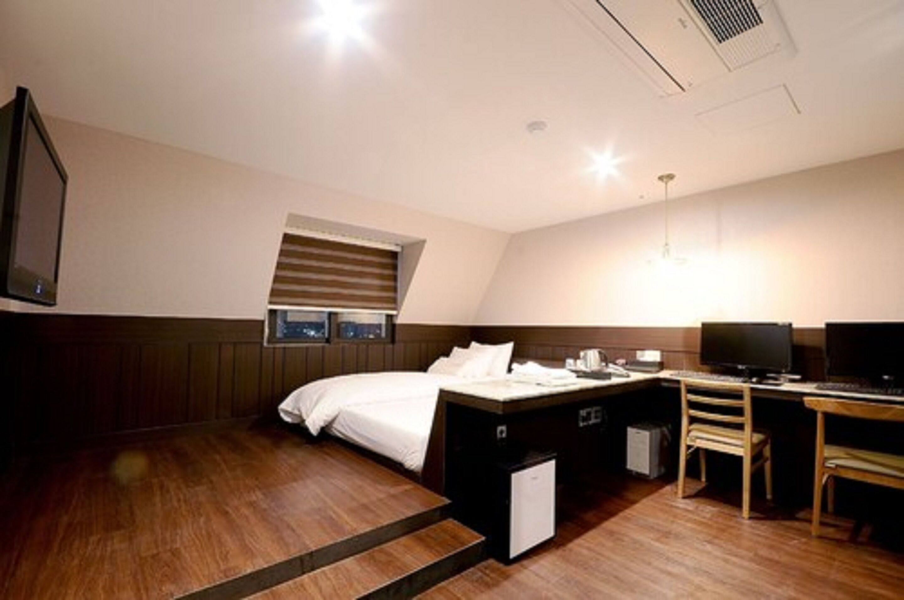 A hotel room with a bed, desk, two monitors, a chair, and a small cabinet.