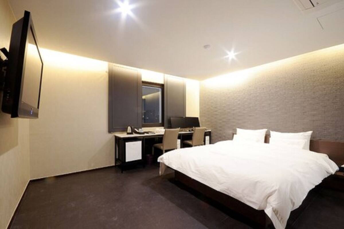 Quarto de hotel com cama, escrivaninha, cadeira e televisão fixada na parede.