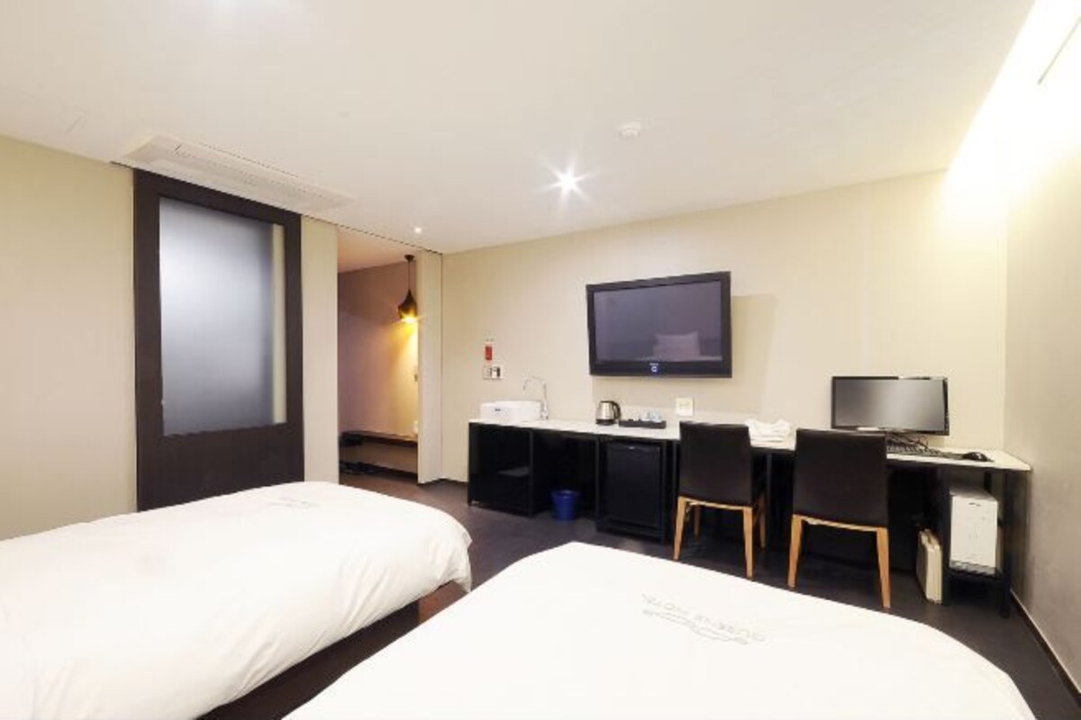 A hotel room with two beds, a desk with a computer, a TV, and a small kitchenette.