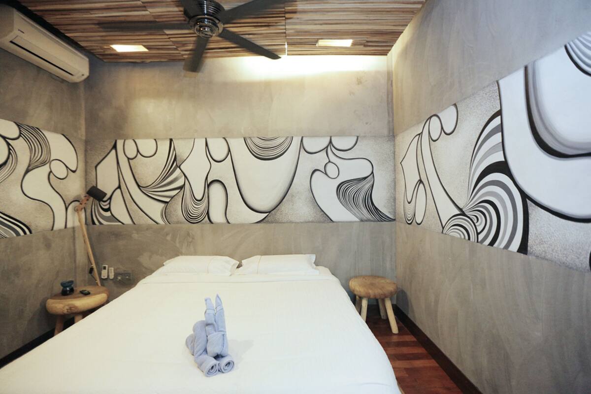 A hotel room with a bed, a ceiling fan, a wall-mounted air conditioner, and abstract wall art.