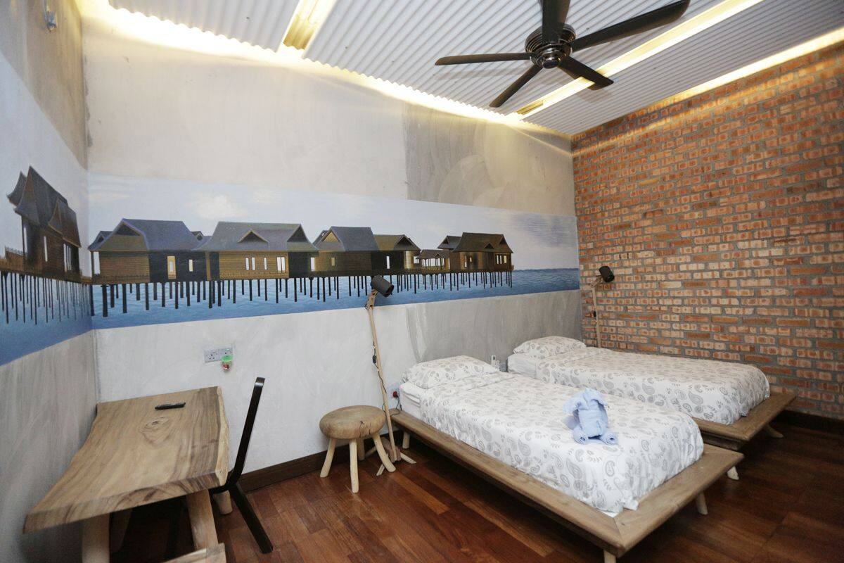 A room with two beds, a wooden table, a chair, a ceiling fan, and a mural of houses on the wall.