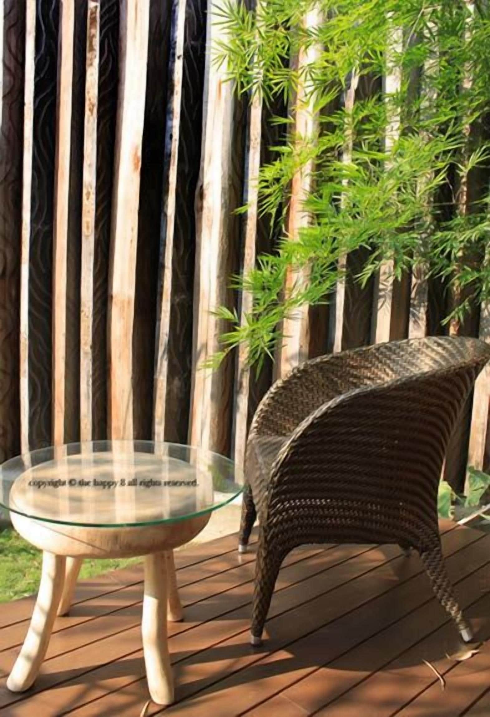 Une table à plateau en verre, accompagnée d’une chaise en osier, posée sur une terrasse en bois, le tout entouré de plantes de bambou.