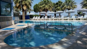 Zona de piscina con sillas de descanso y sombrillas, rodeada de palmeras.