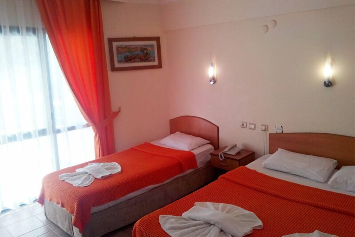 A hotel room with two beds, each with a red blanket and a white towel. There is a painting on the wall, a window with curtains, and a nightstand with a phone.