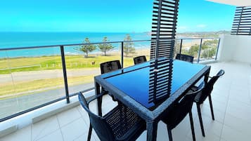 A balcony with a glass railing, a table, and chairs, overlooking a beach and ocean.