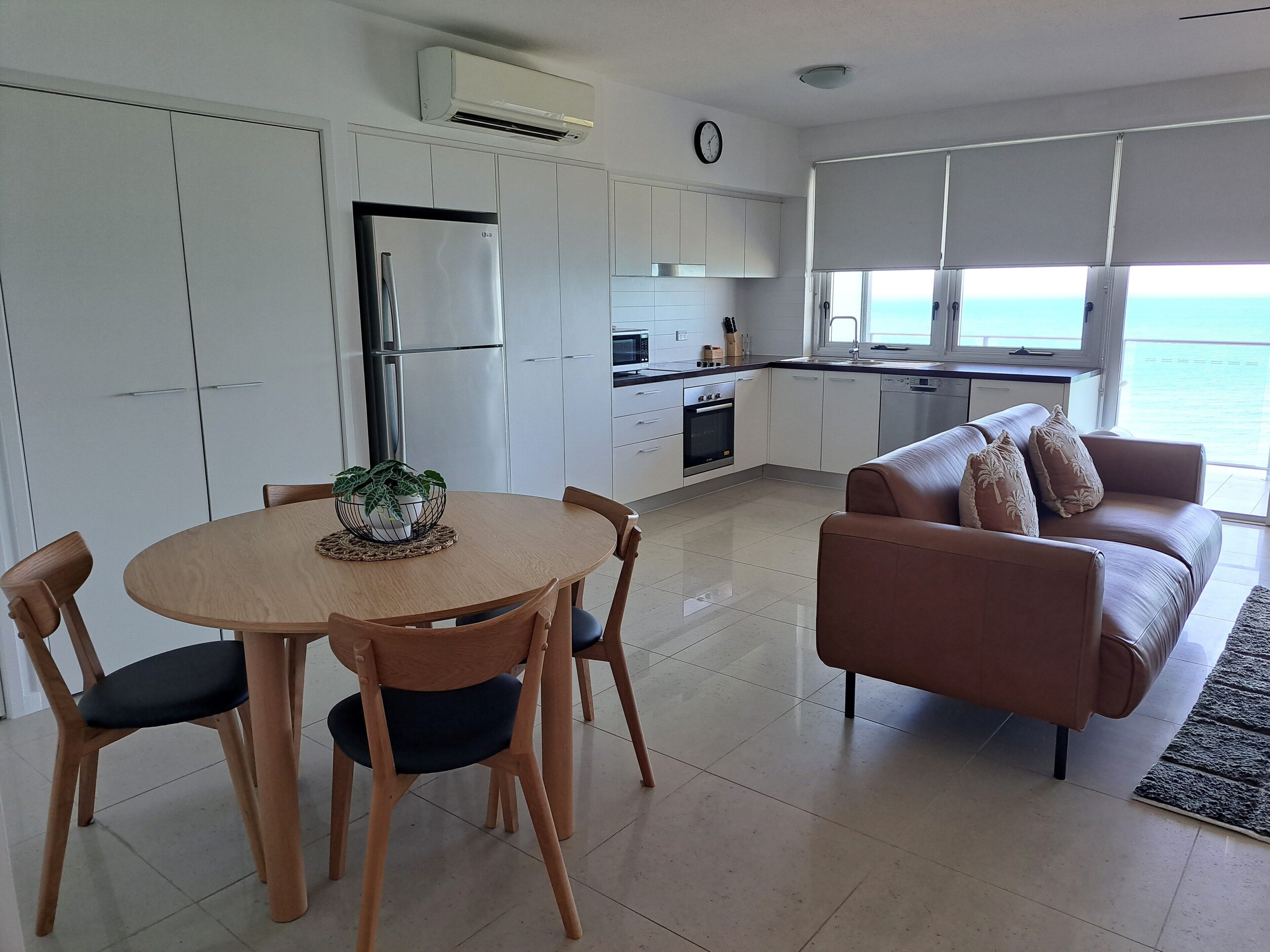 A modern kitchen with white cabinets, a built-in oven, and a refrigerator. There is a dining area with a round table and chairs, and a living room with a brown sofa and a view of the ocean through large windows.