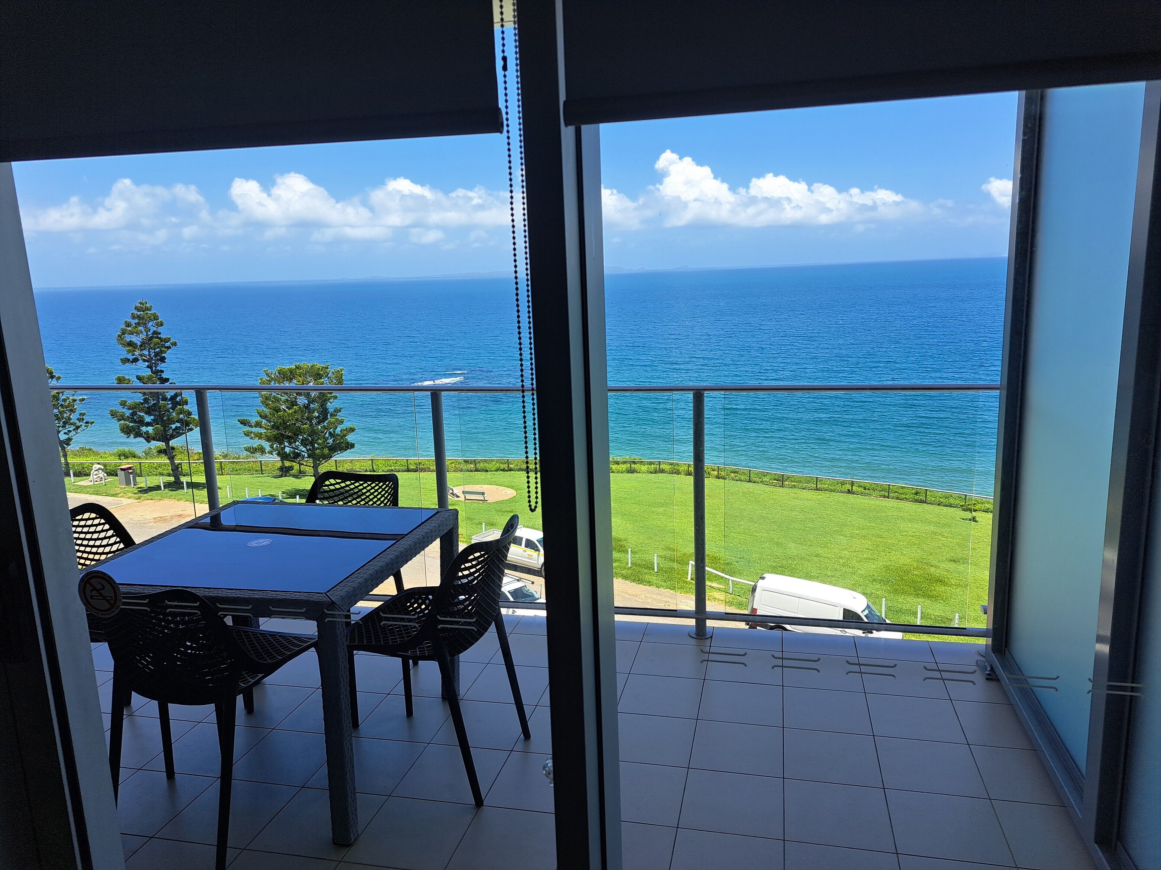 Un balcón con mesa y sillas, con vistas a un espacio verde y al mar.