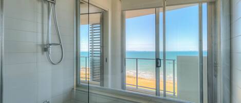 A bathroom with a glass-enclosed shower, a view of the beach, and a balcony with a railing.