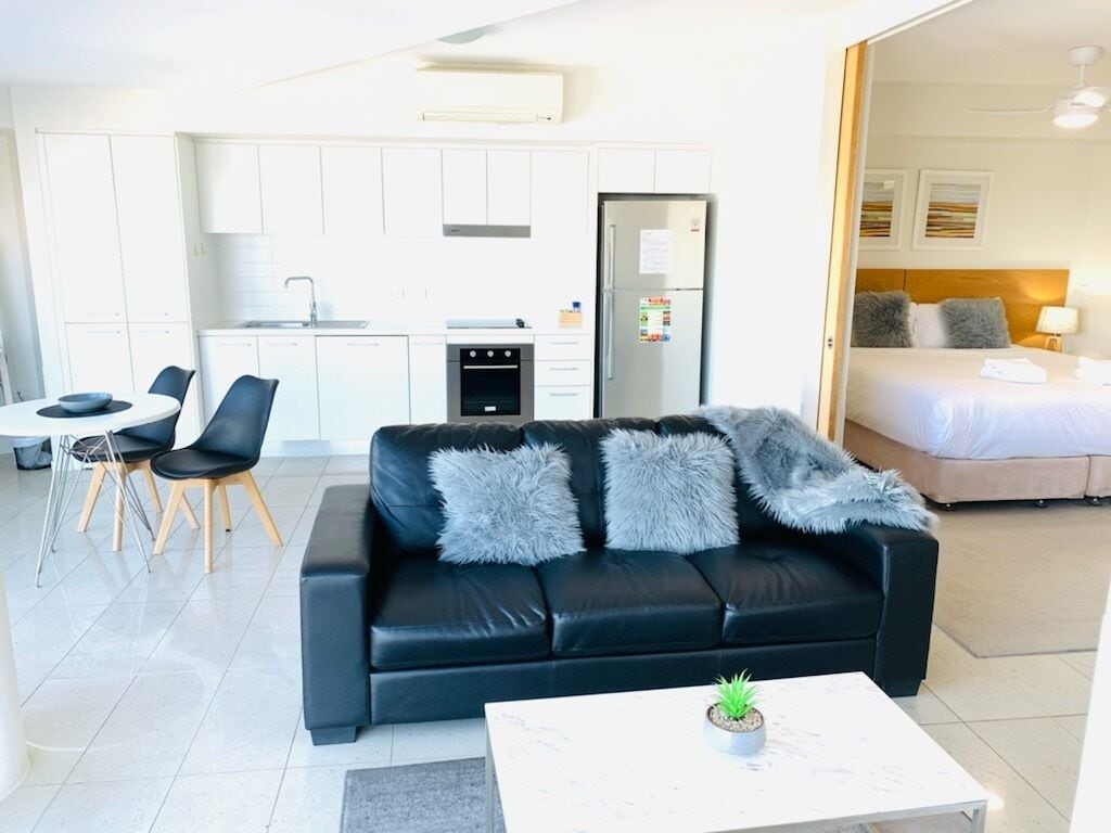 Una sala de estar moderna con un sofá de cuero negro, un comedor con una mesa y sillas blancas, una cocina con gabinetes y electrodomésticos blancos, y un dormitorio con cama y mesita de noche.