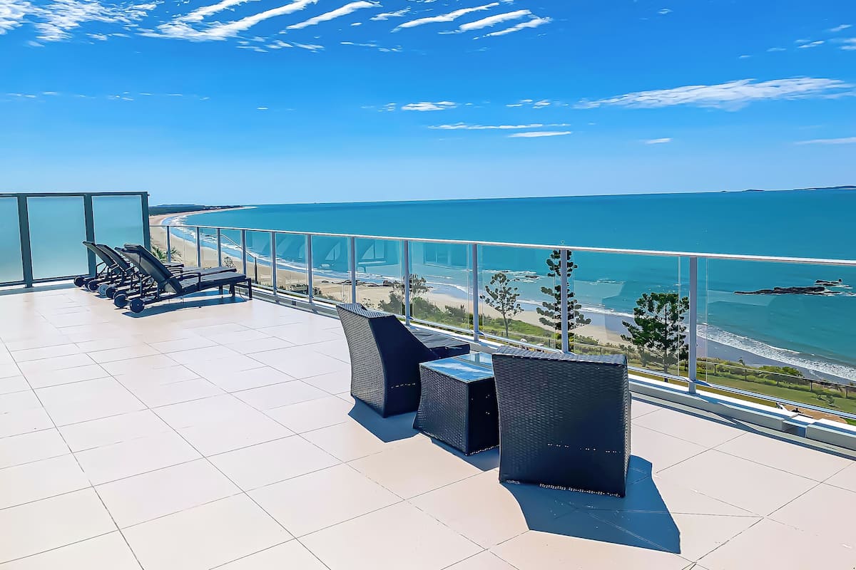 Eine Dachterrasse mit Trainingsgeräten, Sitzgelegenheiten und einem Glasgeländer mit Blick auf Strand und Meer.