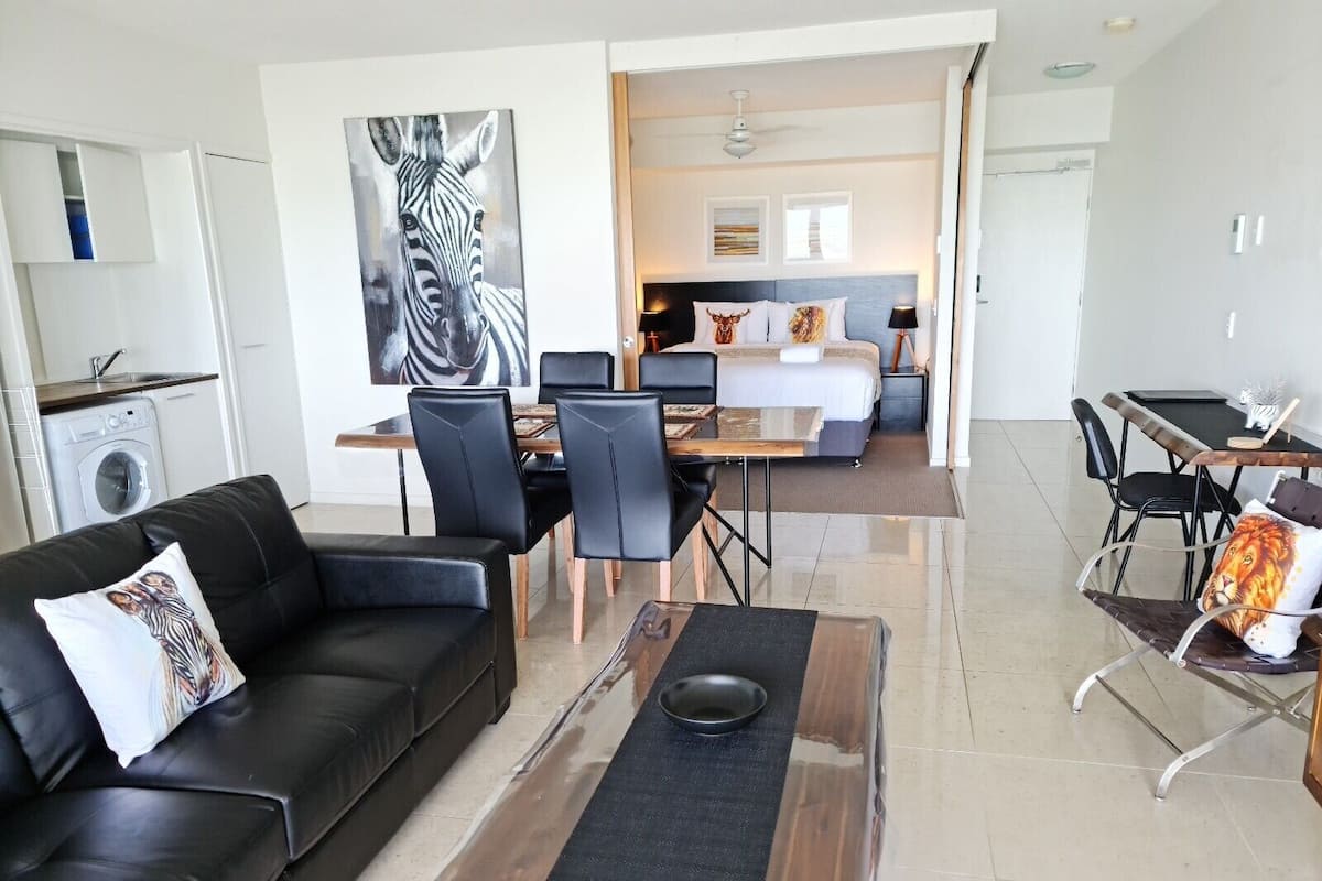 Ein modernes Wohnzimmer mit Sofa, Couchtisch und Essbereich. Im Hintergrund ist eine Küche mit einer Waschmaschine zu sehen.