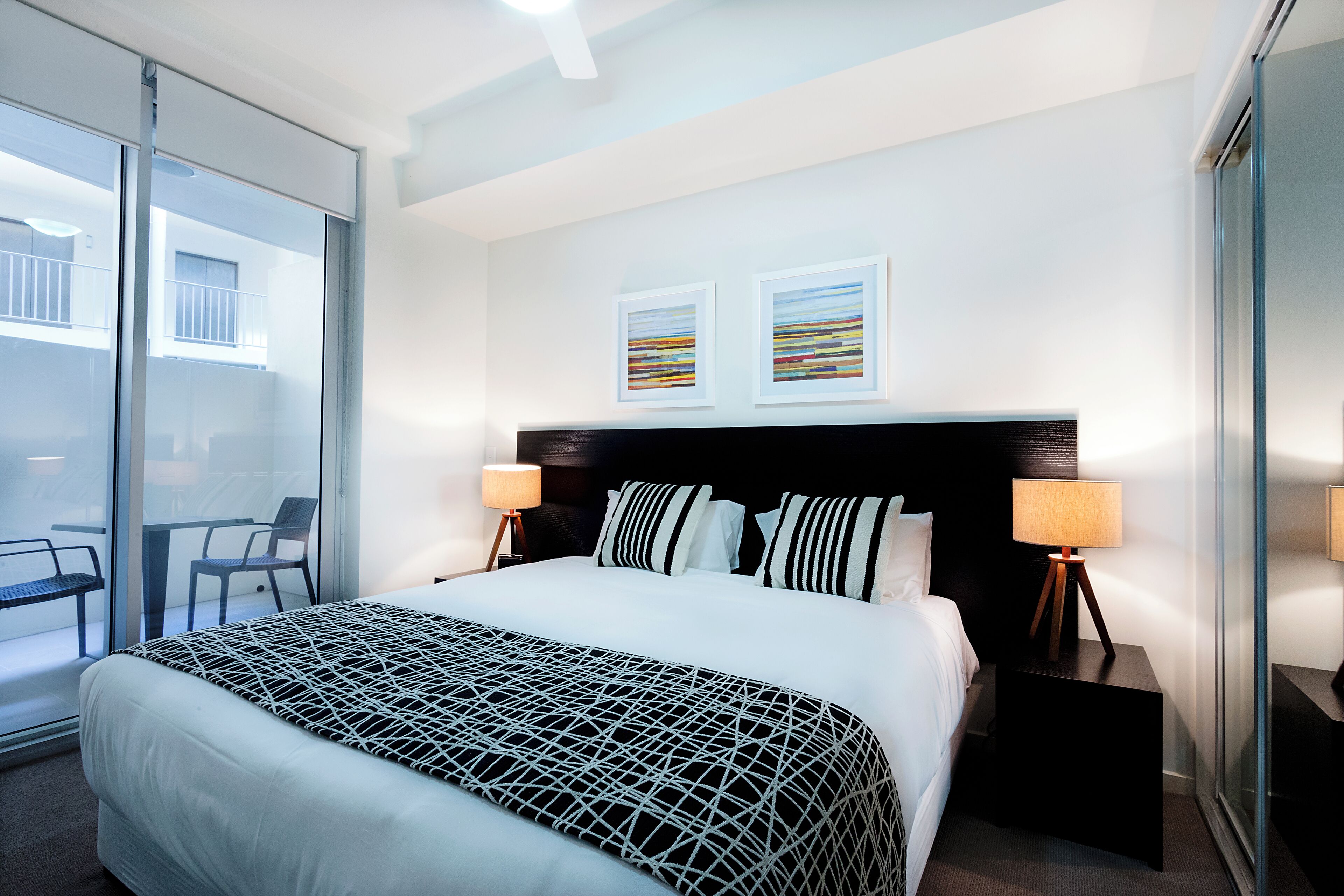 A modern hotel room with a large bed, two bedside tables with lamps, a glass door leading to a balcony, and two framed artworks on the wall.
