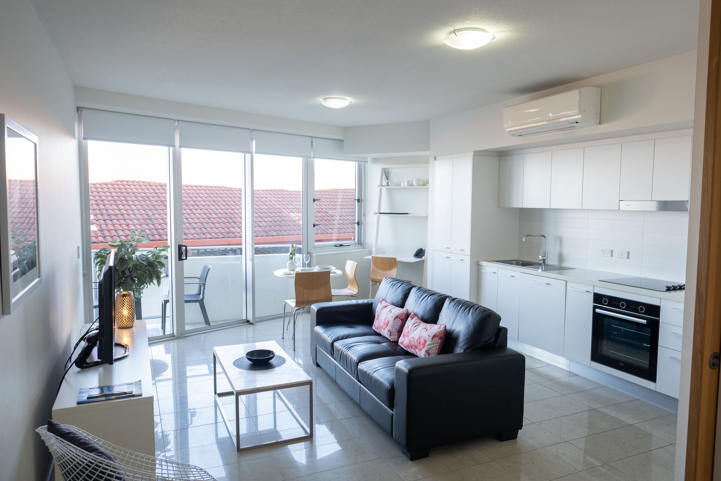 A modern apartment with a living room, dining area, and kitchen. It features a large sliding glass door to the balcony, a black leather sofa, a dining table, and a kitchen with white cabinets and a built-in oven.