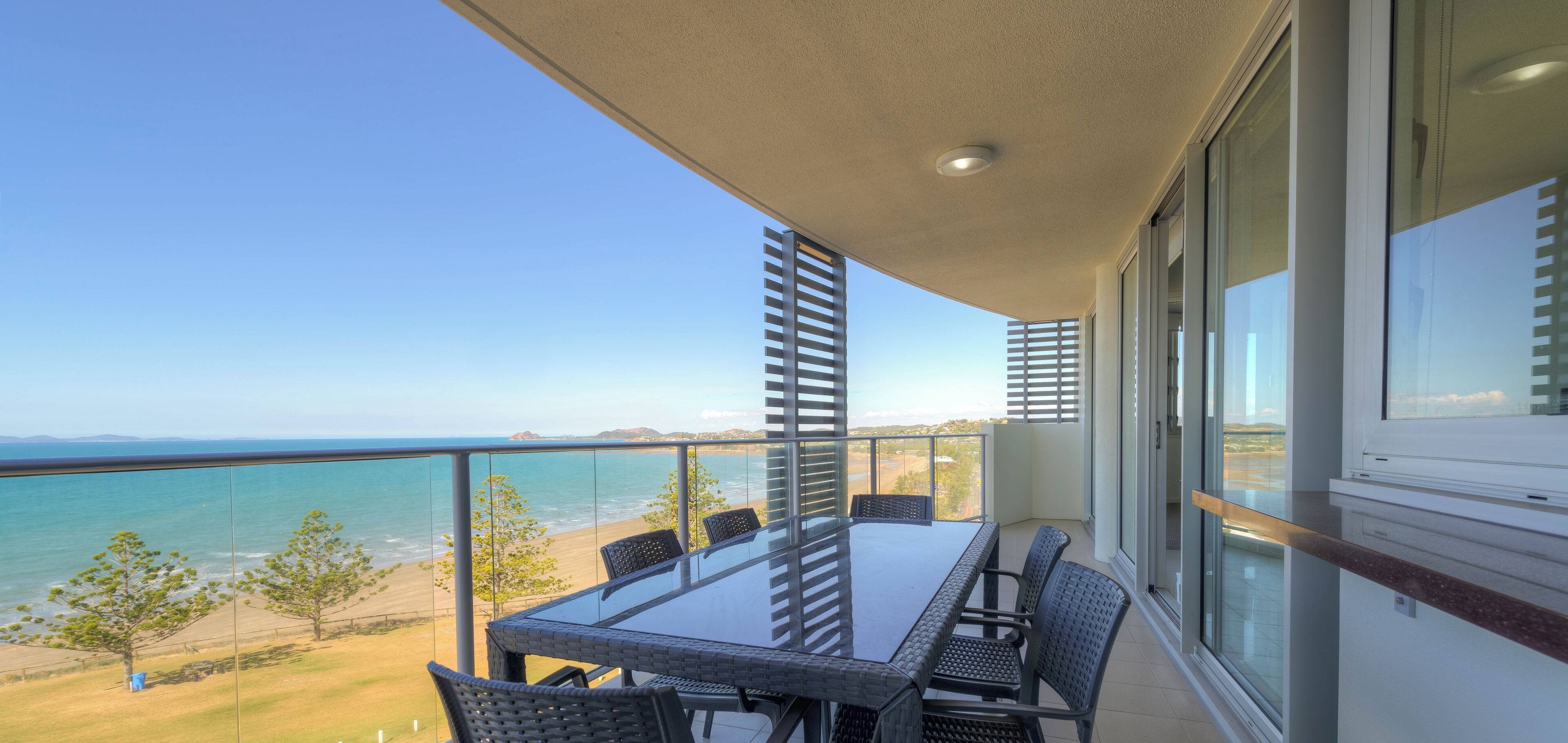 A balcony with a glass table and chairs, overlooking a beach and ocean.