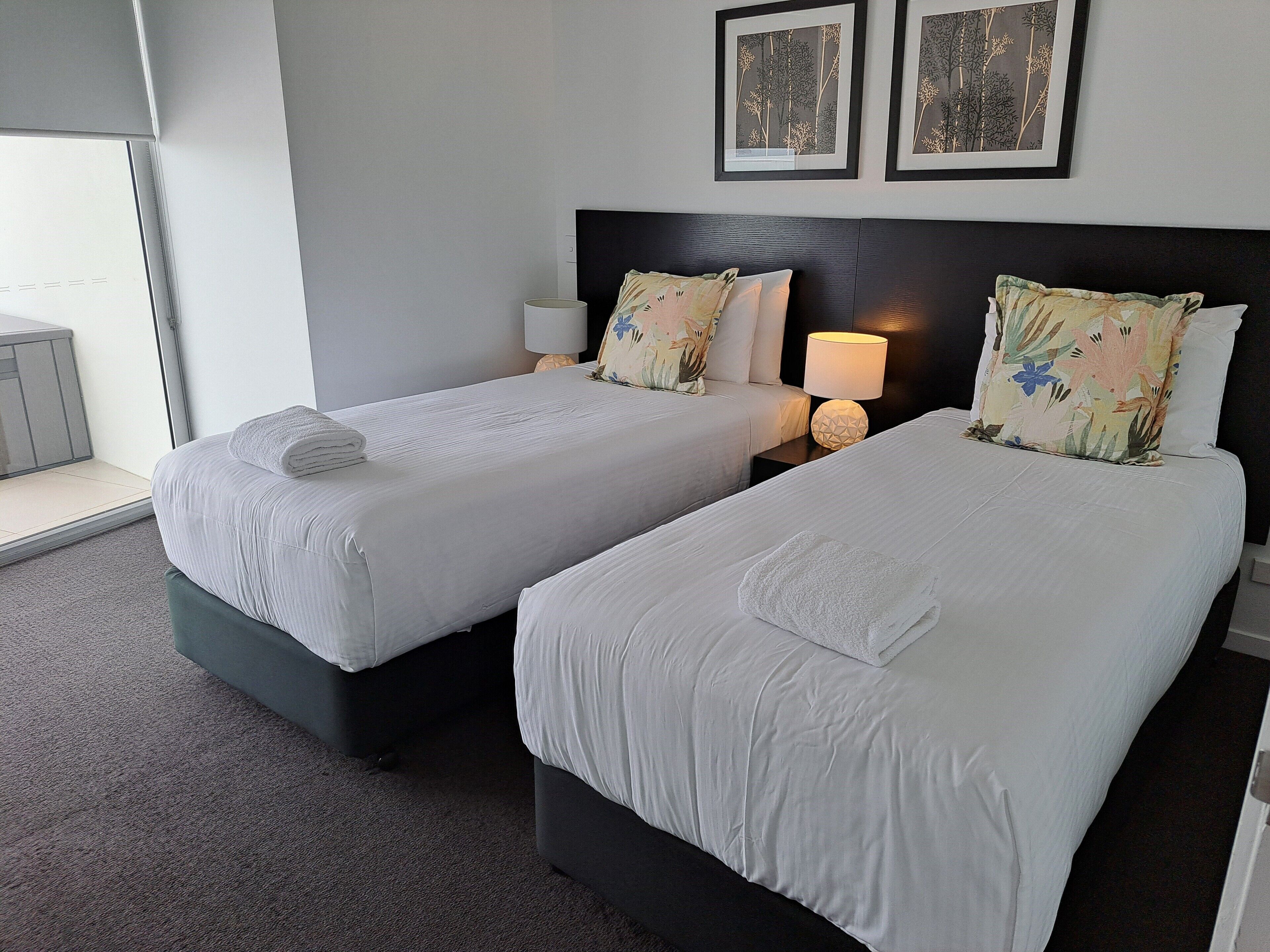 A hotel room with two single beds, a nightstand with a lamp, and two framed botanical prints on the wall.
