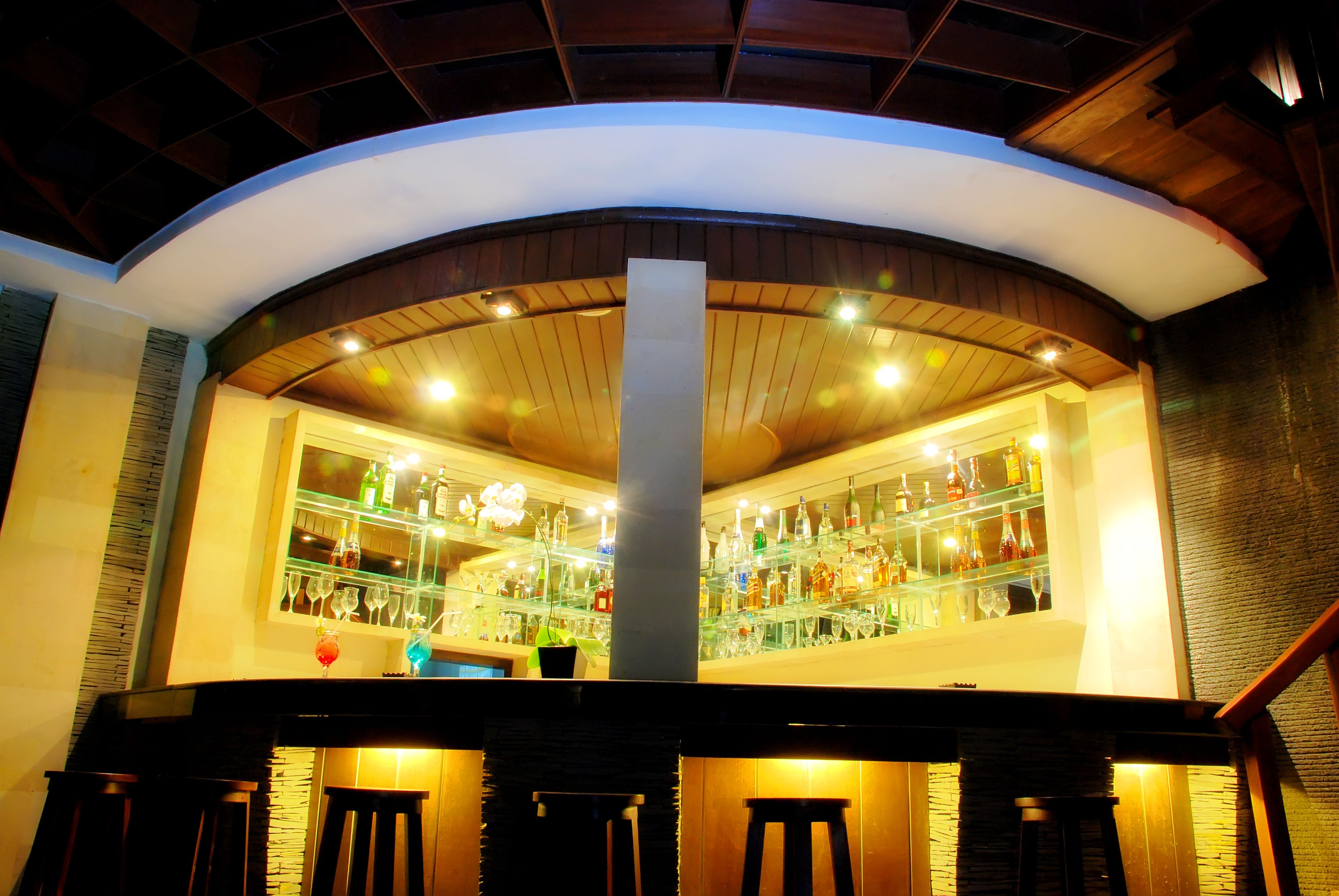 A bar area with a curved ceiling, wooden beams, and a central column. Shelves display various bottles and glasses.