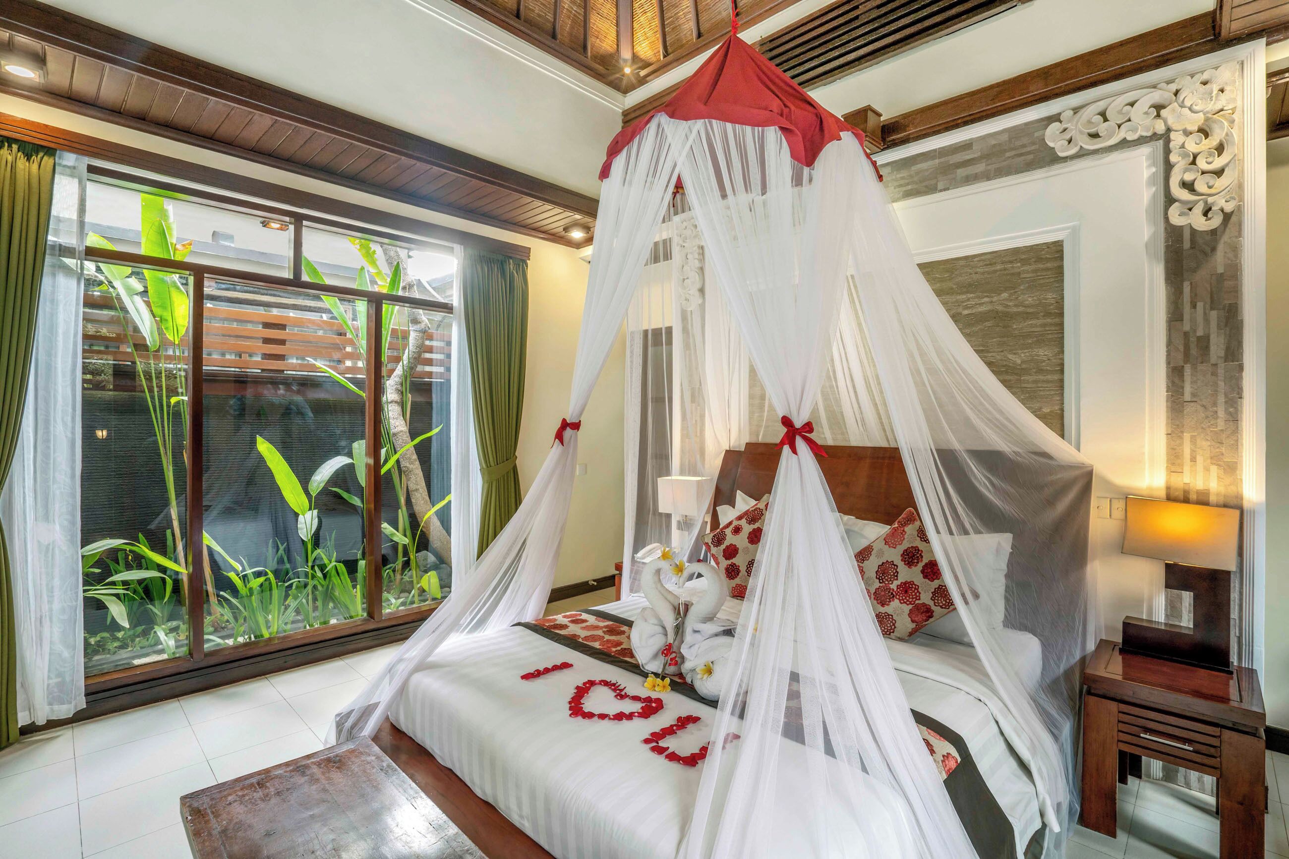 A four-poster canopy bed with a heart-shaped decoration on the bedspread, a nightstand with a lamp, and a view of tropical plants through large windows.