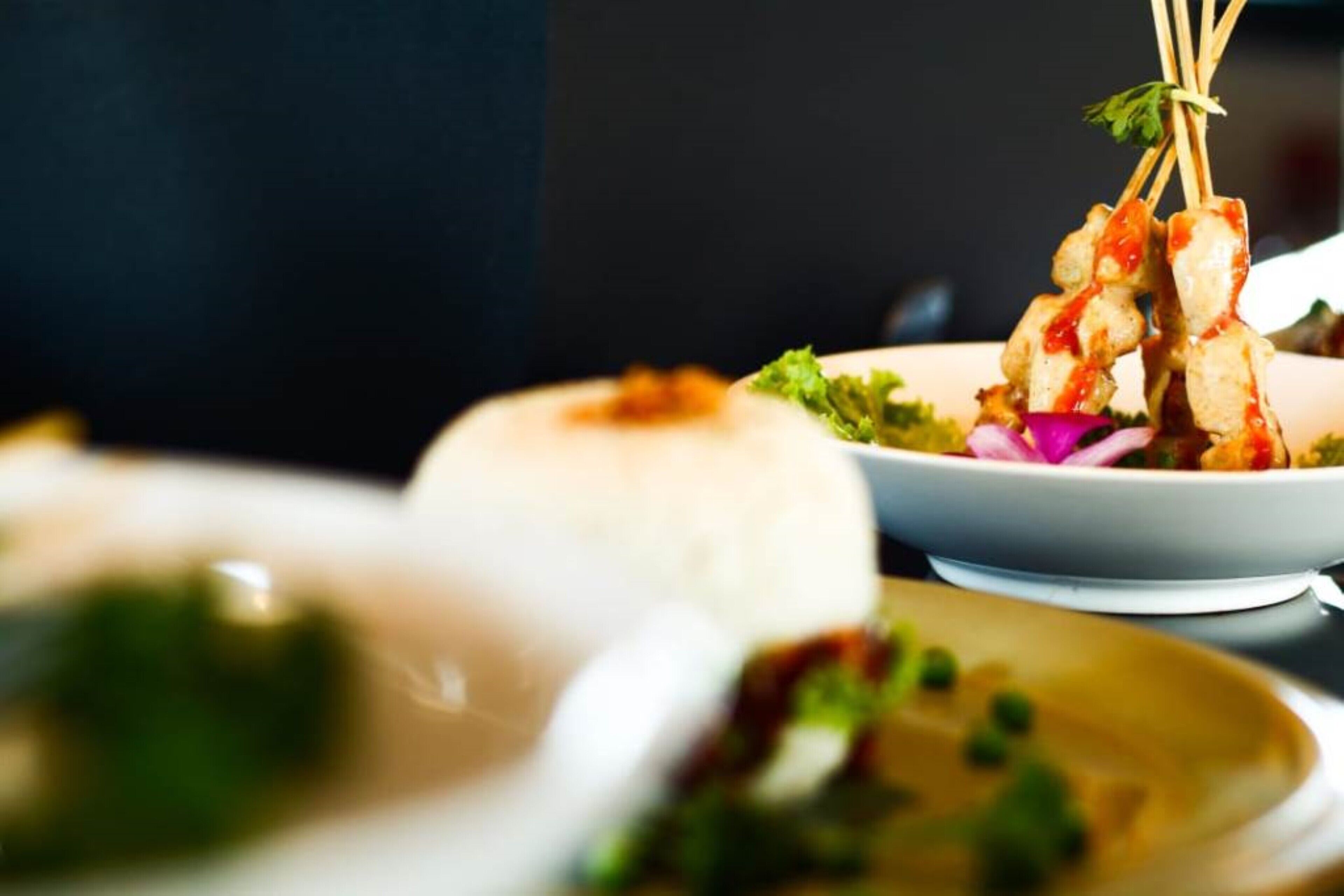 A close-up of a dish with skewered meat and vegetables, a bowl of soup, and a plate with a mound of rice.