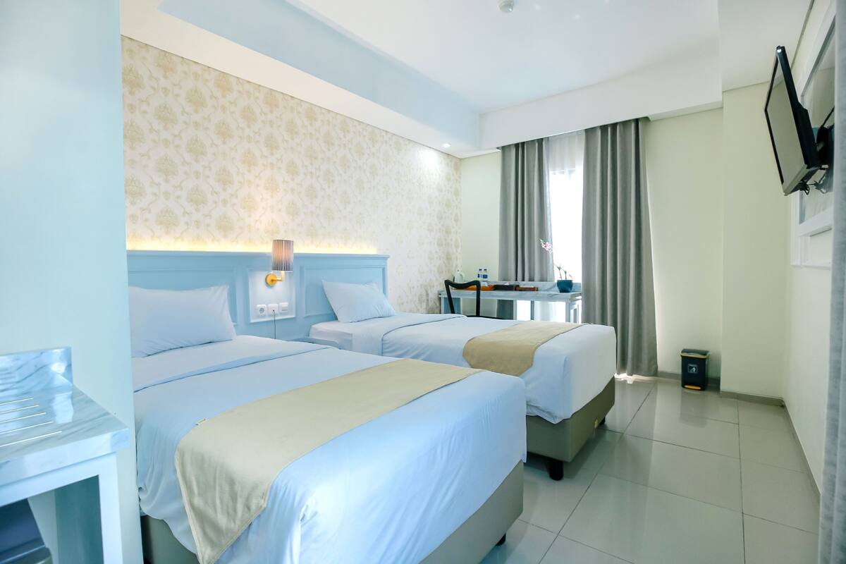A hotel room with two beds, a desk, a chair, a TV mounted on the wall, and a window with curtains.