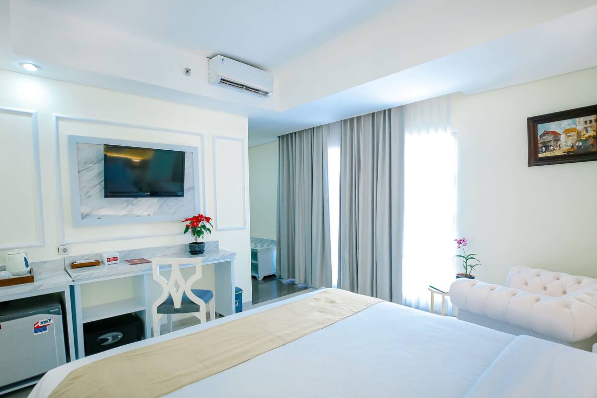 A hotel room with a bed, a desk with a TV, a chair, a sofa, and a window with curtains.