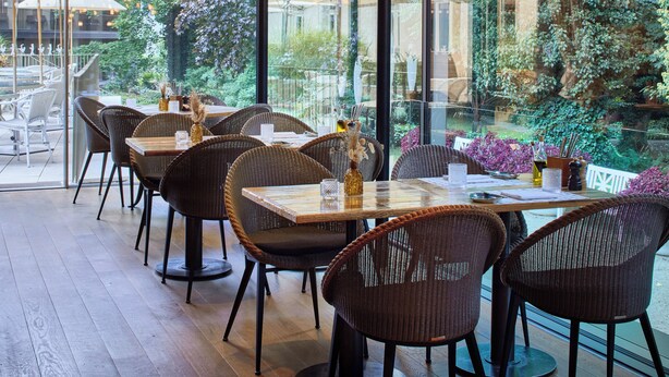 An outdoor dining area with wicker chairs and tables, large umbrellas, and hanging light fixtures. - LA Maison Hotel (Saarlouis)