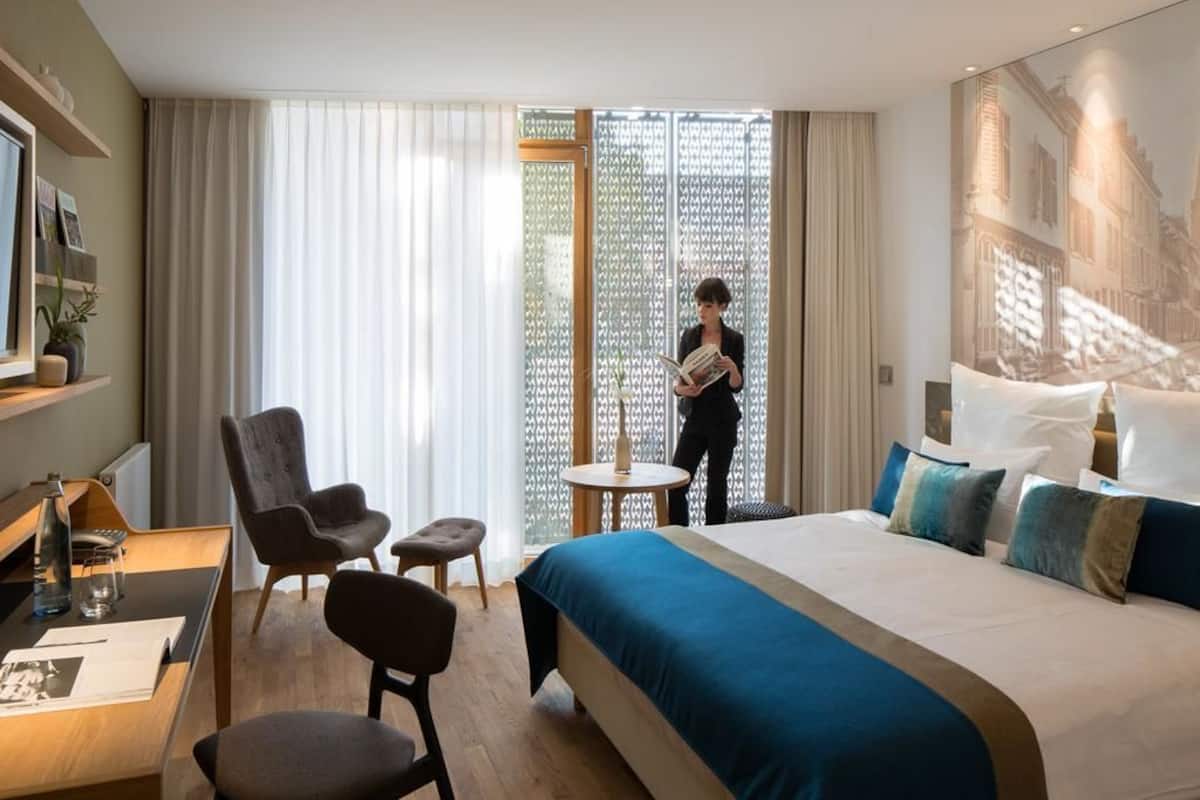A hotel room with a large bed, a desk, two chairs, a small table, and a large window with sheer curtains.