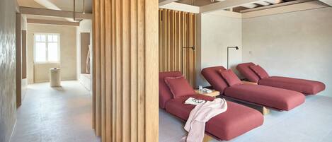 A modern spa room with a wooden partition, two red massage beds, a small table with a glass, and a window providing natural light.