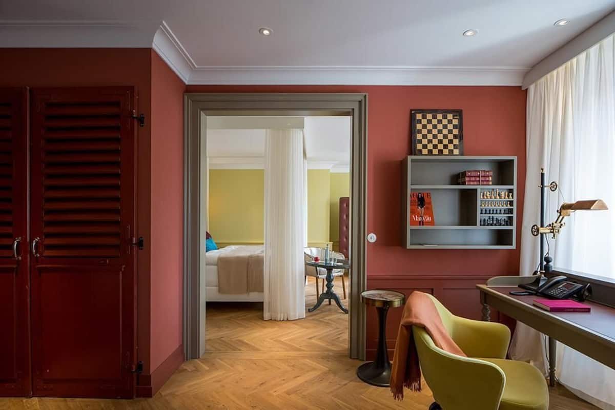 A hotel room with a wooden floor, a bed with white bedding, a desk with a lamp, and a bookshelf.