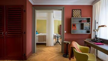Una habitación de hotel con piso de madera, una cama con ropa blanca, un escritorio con lámpara y una estantería.