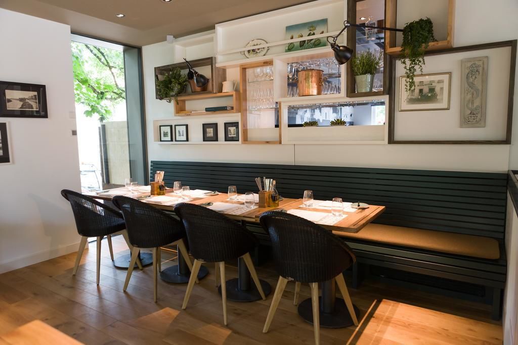A dining area with a wooden table, black chairs, and a bench. Shelves with plants and framed pictures are above the table.