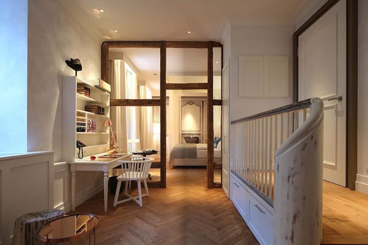 A hallway with a wooden floor, a white desk with a chair, a bed, and a staircase with a white railing.