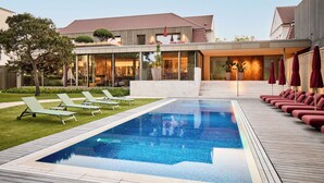 A swimming pool with lounge chairs and umbrellas, surrounded by a wooden deck and a building with large windows. - LA Maison Hotel (Saarlouis)
