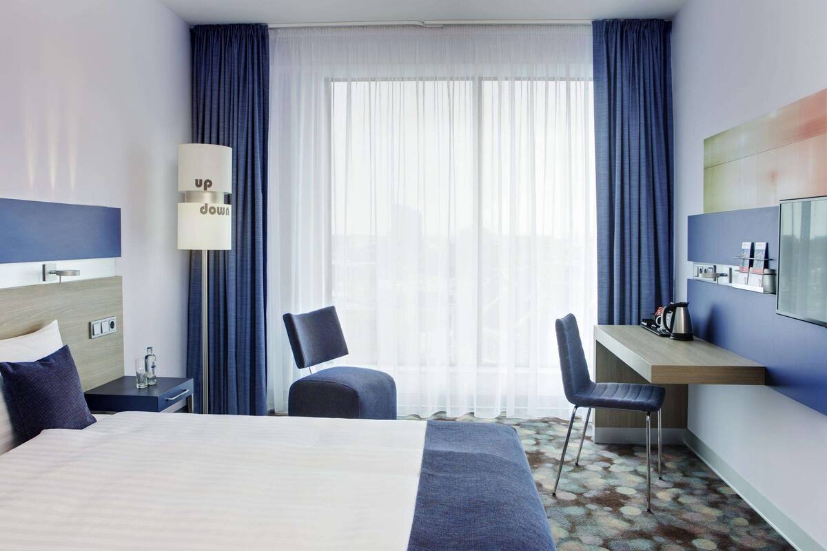 A hotel room with a bed, a desk, a chair, a lamp, and a large window with curtains.