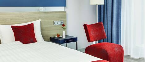 Habitación de hotel con cama, mesita de noche, una silla roja, lámpara y ventana con cortinas.