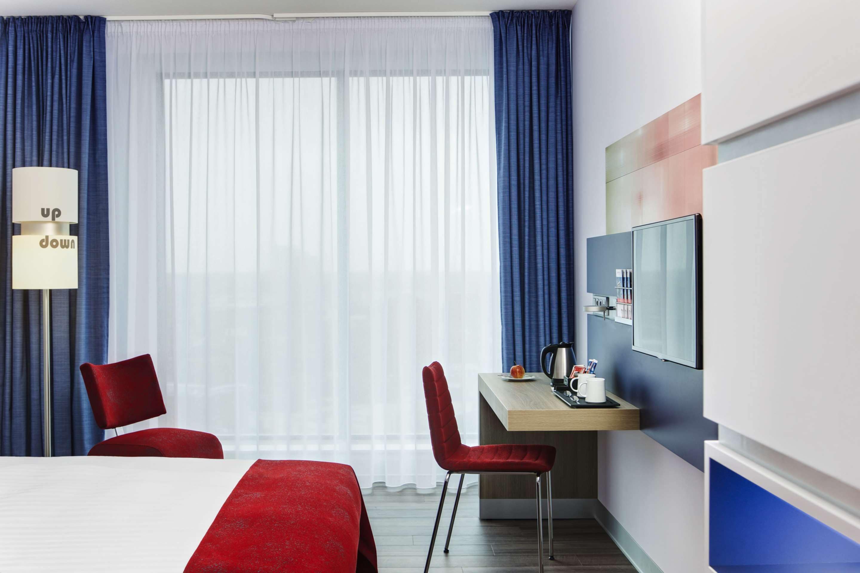 Habitación de hotel con cama, dos sillas rojas, escritorio con cafetera y televisor en la pared.