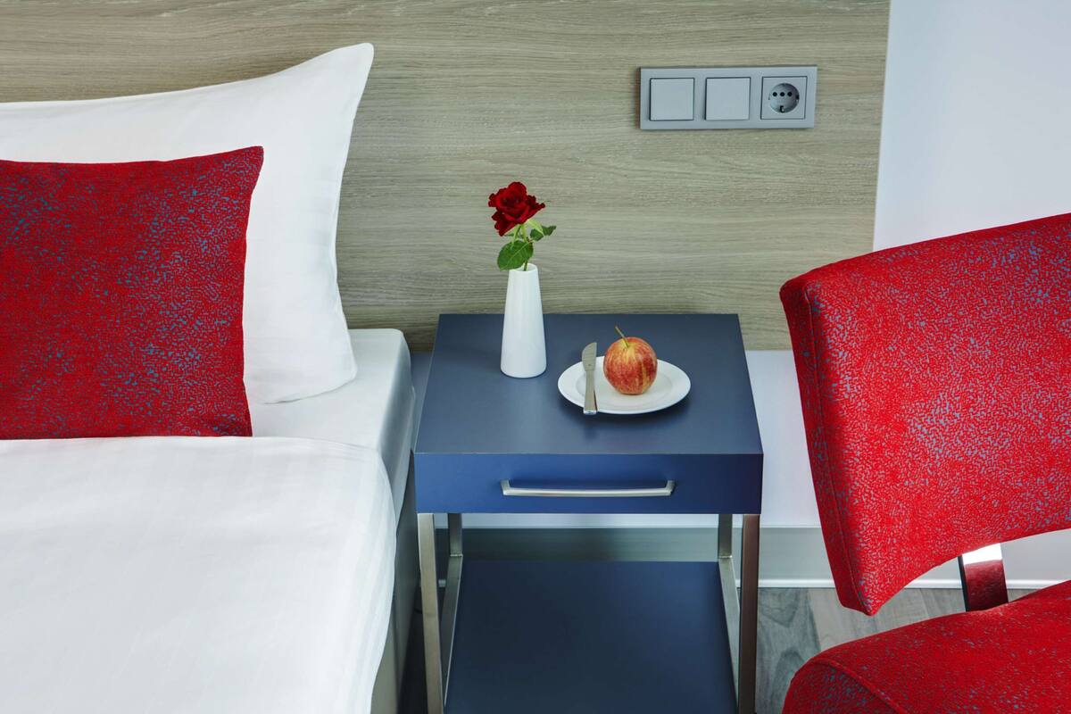 A hotel room with a bed, a nightstand with a vase of flowers and an apple, and red upholstered chairs.